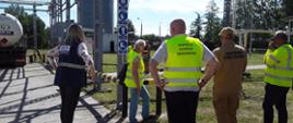 Inspektorzy WIOŚ w Warszawie, przedstawicielka Polski Gaz s.a. i funkcjonariusz państwowej straży pożarnej prowadzą oględziny autocysterny. Pojazd stoi na utwardzonym placu, teren dookoła porośnięty jest trawą. Stoją na nim budynki, drzewa i latarnie.