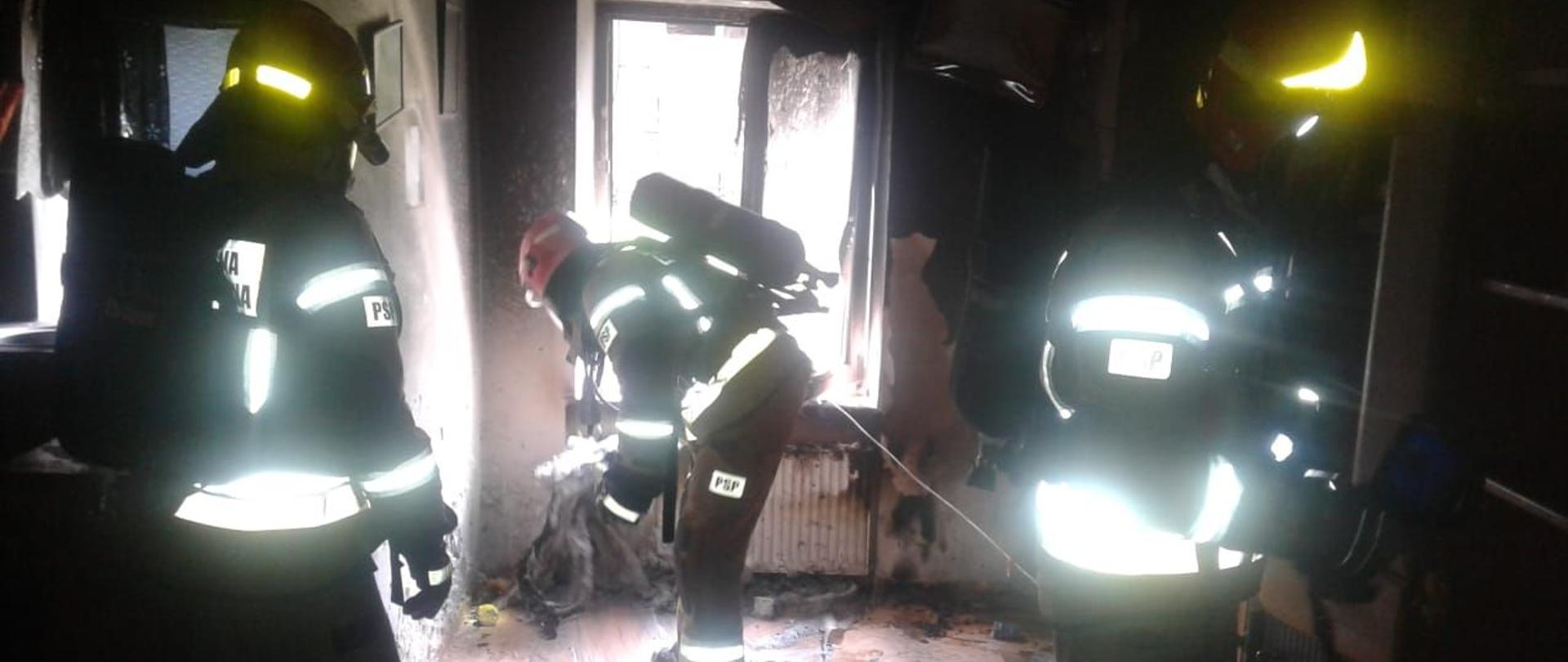 W pomieszczeniu w którym powstał pożar trzech strażaków usuwa spalone elementy wyposażenia na zewnątrz przez okno.