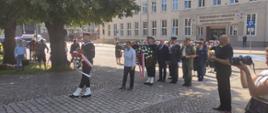 Żołnierz Marynarki Wojennej trzyma wiązankę, za nim idą zaproszeni goście oraz przedstawiciele służb mundurowych.