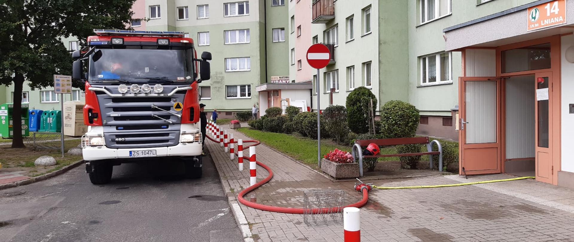 Samochód ratowniczo gaśniczy przed klatką schodową budynku mieszkalnego, z rozwinięta linią gaśniczą do budynku.