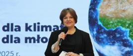 Wiceminister Zioło-Pużuk stoi na tle wielkiego banera z napisem Młodzi dla klimatu - klimat dla młodych, mówi do trzymanego w ręku mikrofonu, w ręku trzyma kartkę papieru.
