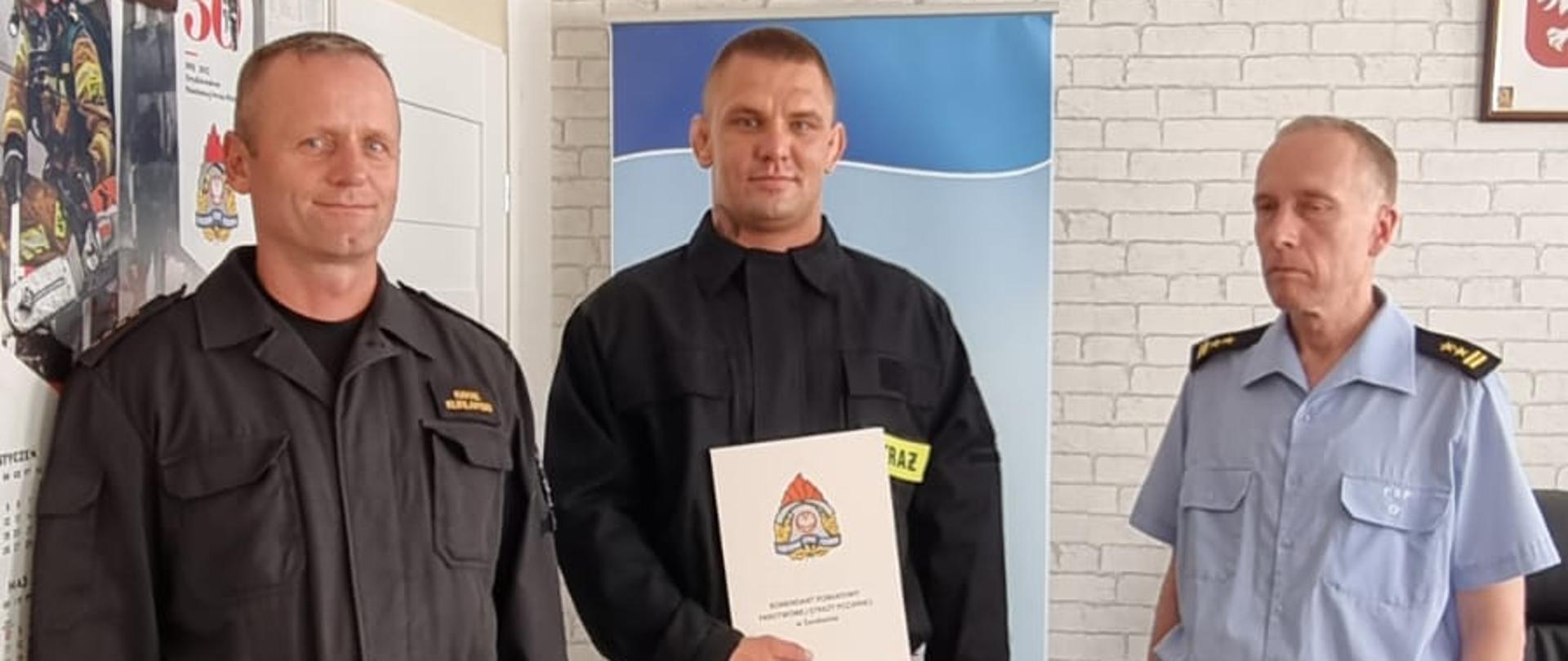 Zdjęcie przedstawia trzech mężczyzn ubranych w czarne mundury służbowe, jeden z nich trzyma w rękach dokumenty. Za nimi widnieje baner z logiem PSP oraz napisem: "Komenda Powiatowa Państwowej Straży Pożarnej w Świdwinie". Za banerem widzimy ścianę z tapetą w szare cegły. Na ścianie wisi Godło Rzeczypospolitej Polskiej. Zdjęcie wykonano w gabinecie Komendanta Powiatowego PSP w Świdwinie.