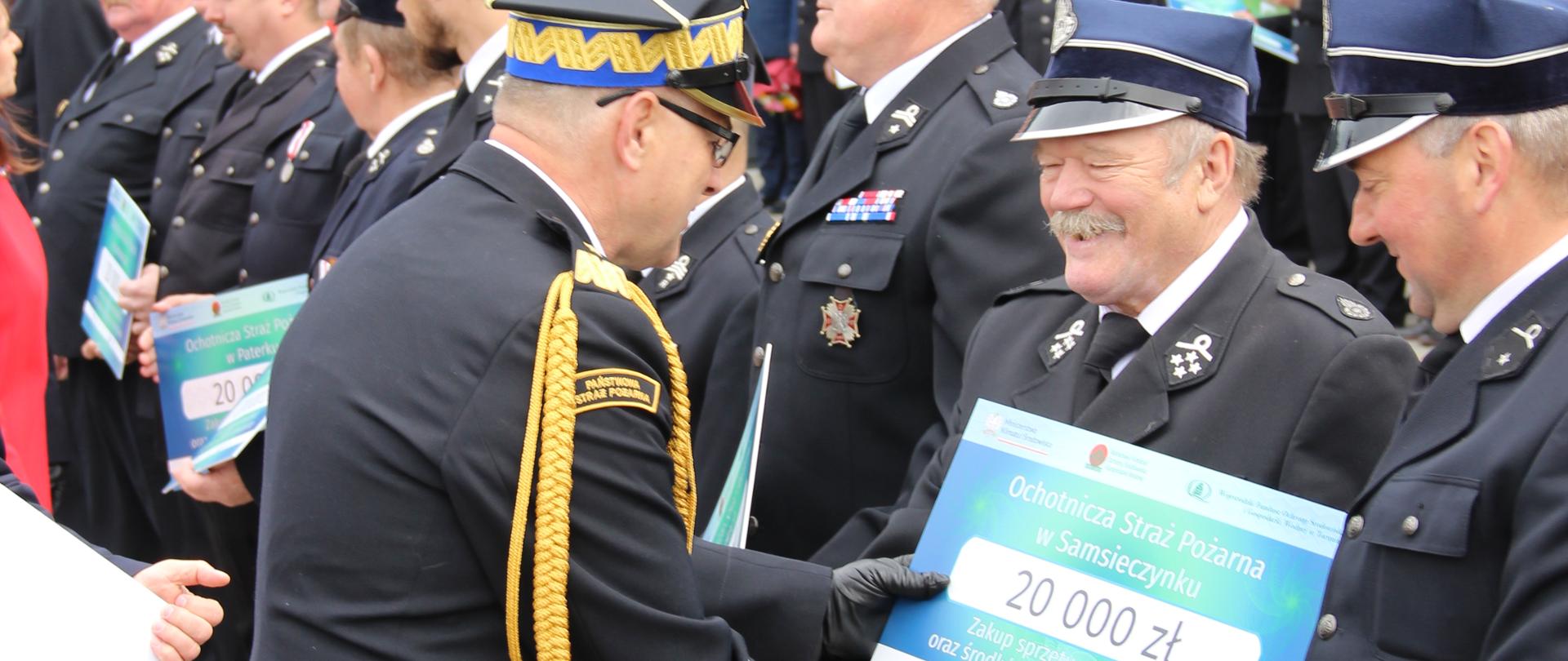 Komendant wojewódzki w mundurze generalskim wręcza voucher druhowi OSP. Druh stoi w szeregu, za szeregiem pododział strażaków w mundurach wyjściowych. W tle budynek strażnicy.