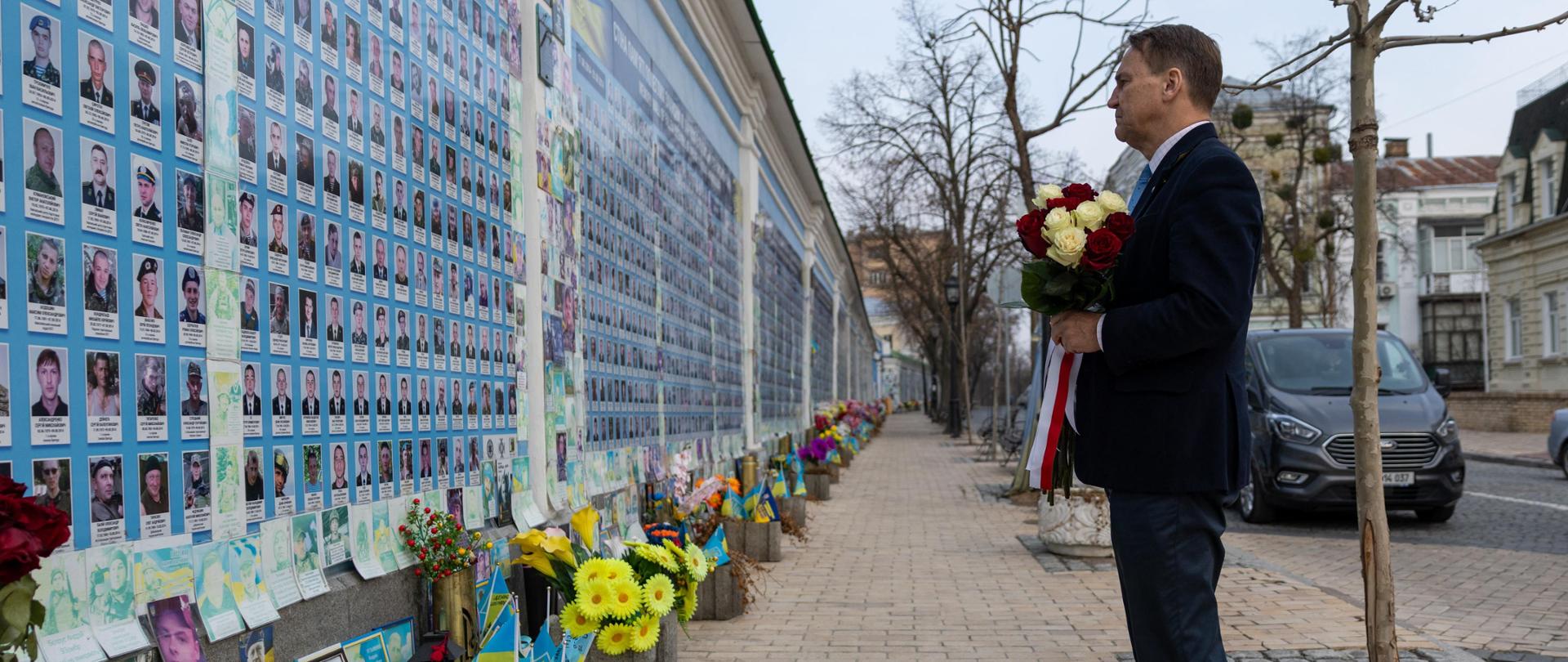 Na zdjęciu jest mężczyzna w garniturze. W rękach trzyma bukiet białych i czerwonych róż. Jest zwrócony w kierunku ściany na której są zdjęcia wielu osób. Pod ścianą są kwiaty. 