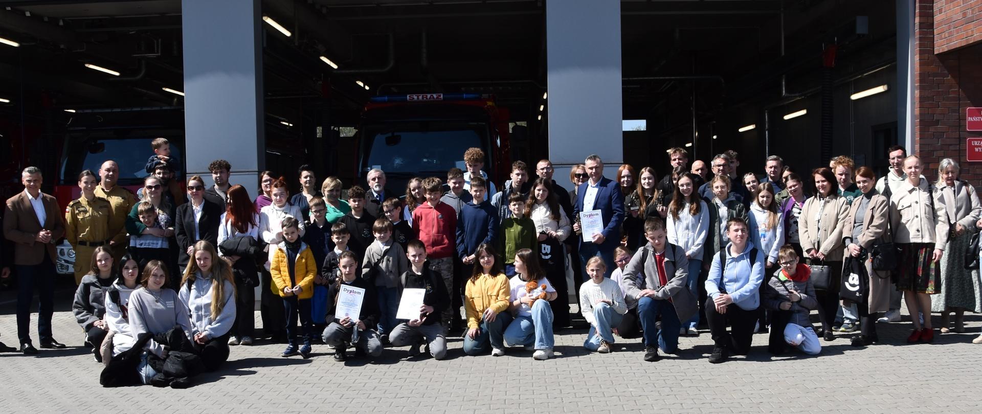 Na zdjęciu uczestnicy eliminacji i zamroczeni goście zgromadzeni przed budynkiem komendy. W budynku otwarte bramy, widać samochody strażackie zaparkowane w garażu. 