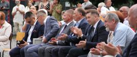 Na dworze pod drzewami na ustawionych w rzędy krzesłach siedzą ludzie, w pierwszym rzędzie minister Czarnek.