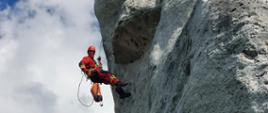 Na zdjęciu widać ratowników wysokościowych w kombinezonach wysokościowych w kolorze czerwonym. Zdjęcie wykonane w górach, w ciągu dnia. Skały w kolorze jasnoszarym, częściowo pokryte mchem.