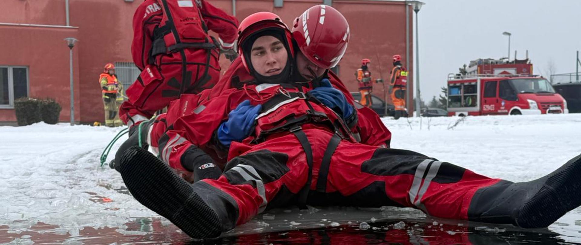Dwaj ratownicy w czerwonych kombinezonach leżą na lodzie przy przeręblu, jeden asekurując drugiego; na pierwszym planie widać wodę i krę lodową, w tle budynek oraz wozy i strażaków biorących udział w ćwiczeniach.
