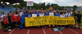 zwycięska drużyna oraz strażacy i druhowie pozują do zdjęcia na stadionie, przed sobą trzymają baner z odręcznym napisem mdp osp kunice mistrzem województwa