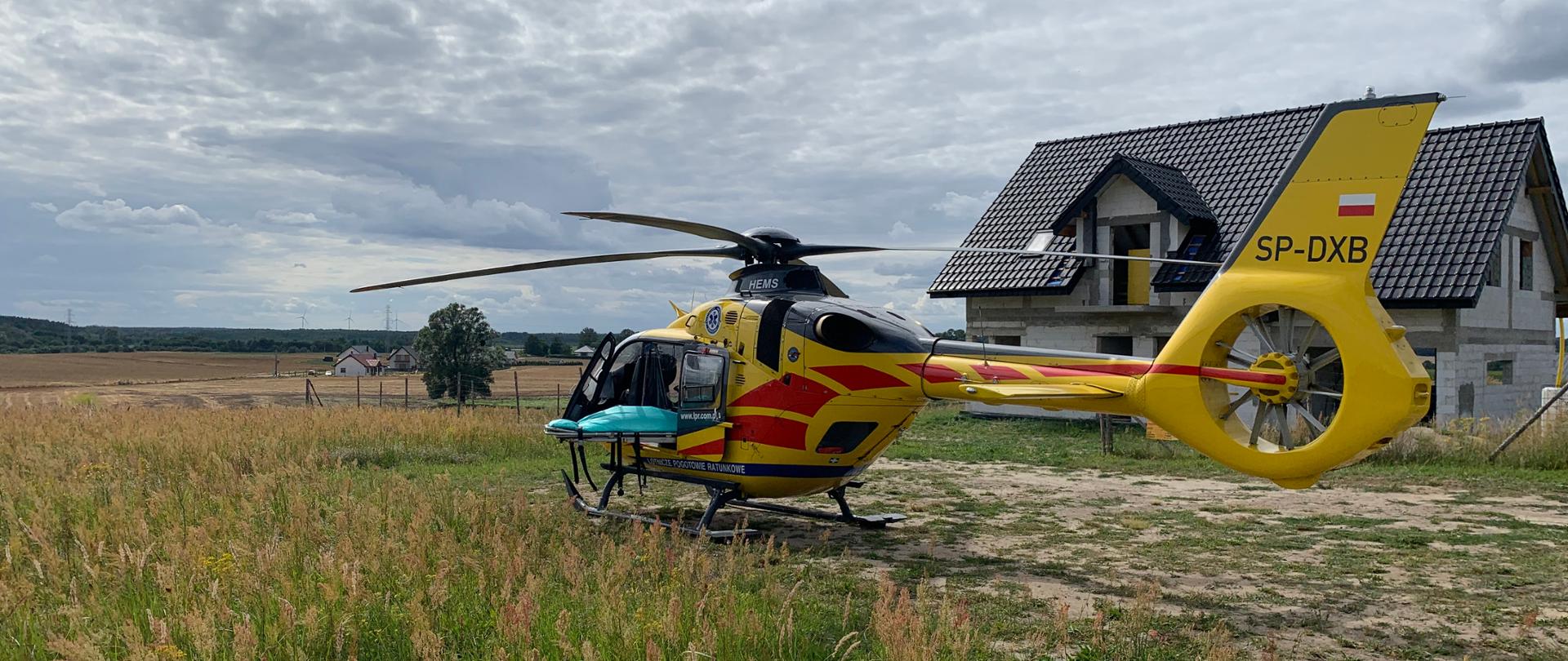 Na fotografii znajduje się śmigłowiec Lotniczego Pogotowia Ratunkowego, który wylądował na podwórku przed domem. Śmigłowiec jest koloru żółtego z czerwonymi elementami. Przednie lewe oraz środkowe lewe drzwi są otwarte. Ze środkowych drzwi wystają nosze medyczne. Po prawej stronie, za śmigłowcem znajduje się dom w trakcie budowy, który ma czarny dach. W oddali znajduje się pole, domki oraz las.