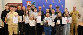 Grupa dzieci i młodzieży stoi w sali konferencyjnej wraz z kilkoma dorosłymi, w tym strażakami. Uczestnicy trzymają dyplomy oraz pudełka z czujnikami dymu, pozując do wspólnego zdjęcia na tle niebieskiej ścianki z herbami.