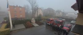 Strażacy w dwóch rzędach stoją przed samochodami pożarniczymi poczet flagowy wiesza flagę Polski na maszt.