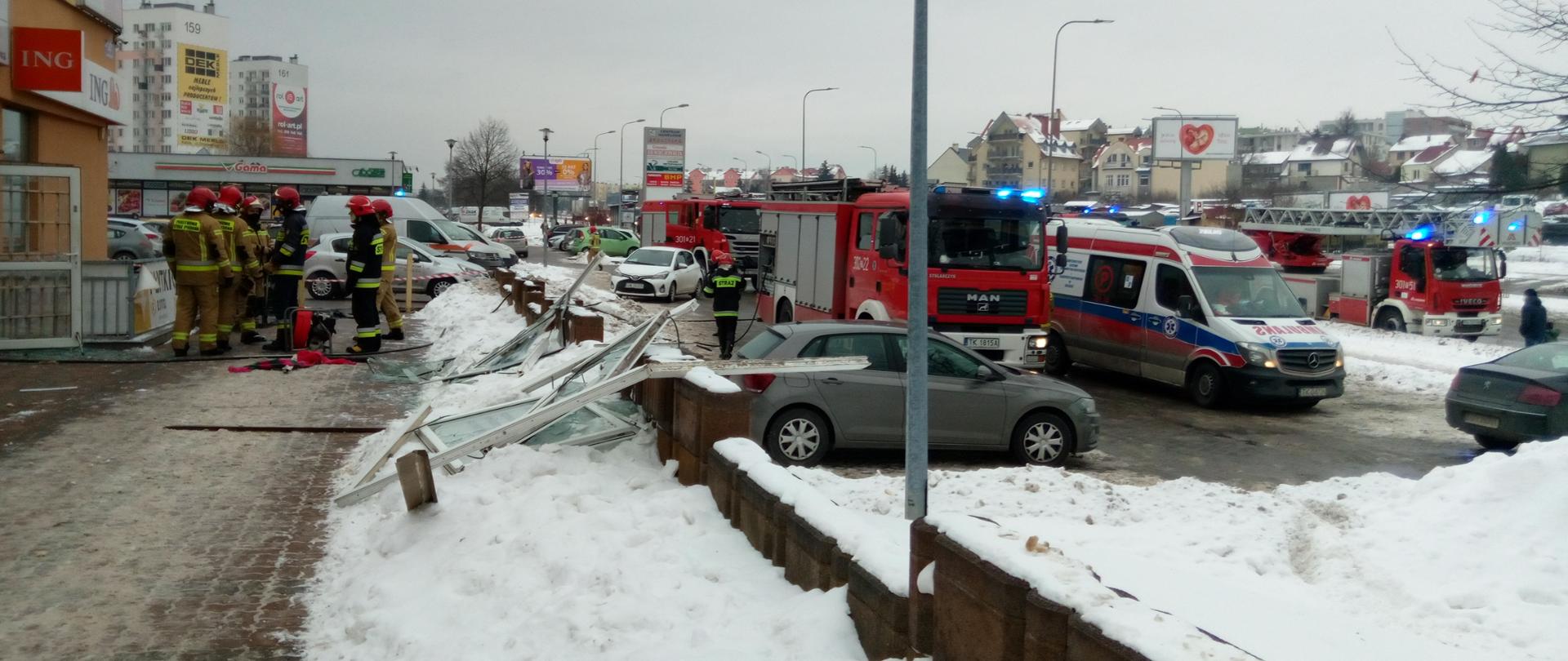 Na zdjęciu widać grupę strażaków stojącą przed budynkiem pralni.
Na chodniku znajduje się wybite okno. Zaraz za chodnikiem widać samochód strażacki, karetkę pogotowia oraz pozostałe samochody osobowe znajdujące się na parkingu.
