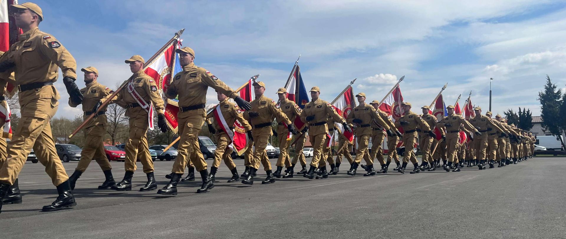 Maszerująca kolumna strażaków w piaskowych mundurach służbowych na asfalcie. Wielu z nich niesie sztandary, w tym polską flagę. Nad nimi błękitne niebo z chmurami, w tle widać zaparkowane samochody.