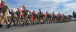 Maszerująca kolumna strażaków w piaskowych mundurach służbowych na asfalcie. Wielu z nich niesie sztandary, w tym polską flagę. Nad nimi błękitne niebo z chmurami, w tle widać zaparkowane samochody.