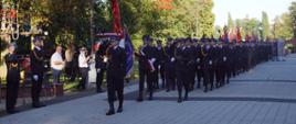 Zdjęcie przedstawia przemarsz defilady z okazji 70-lecia zawodowego pożarnictwa w Zgierzu. Defilada przechodzi przez deptak w parku miejskim w Zgierzu.