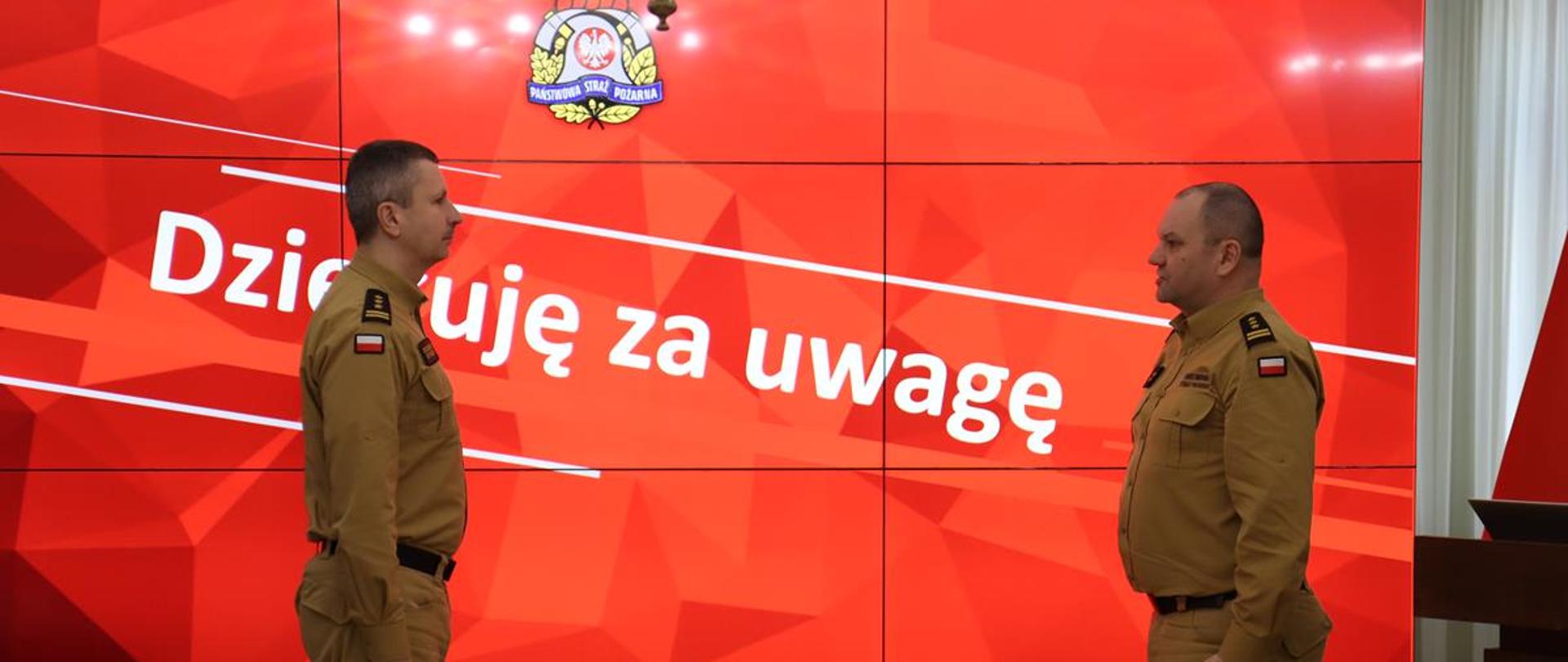 Na zdjęciu dwóch mężczyzn w mundurach Państwowej Straży Pożarnej stoi naprzeciwko siebie w oficjalnej postawie. W tle znajduje się duży ekran z czerwonym tłem i białym napisem „Dziękuję za uwagę”, a nad nim widnieje logo PSP. Scena wygląda na zakończenie oficjalnego wystąpienia lub uroczystości służbowej.