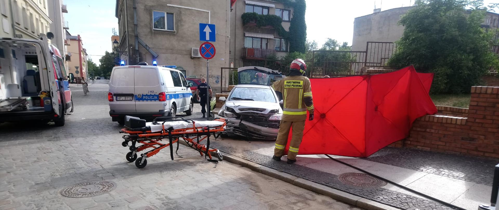 Wpadek z udziałem samochodu osobowego na ul. Polskiej w Brzegu - zdjęcie przedstawia uszkodzony samochód osobowy, z prawej strony widać strażaka trzymającego czerwony parawan z lewej strony stoi karetka pogotowia oraz radiowóz policji