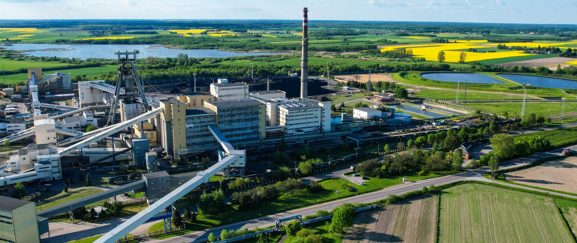 Widok z lotu ptaka na kompleks przemysłowy kopalni wraz z szybem, zabudowaniami technicznymi i infrastrukturą transportową. Wokół zakładu widoczne są tereny zielone, zbiorniki wodne oraz rozległe pola uprawne.