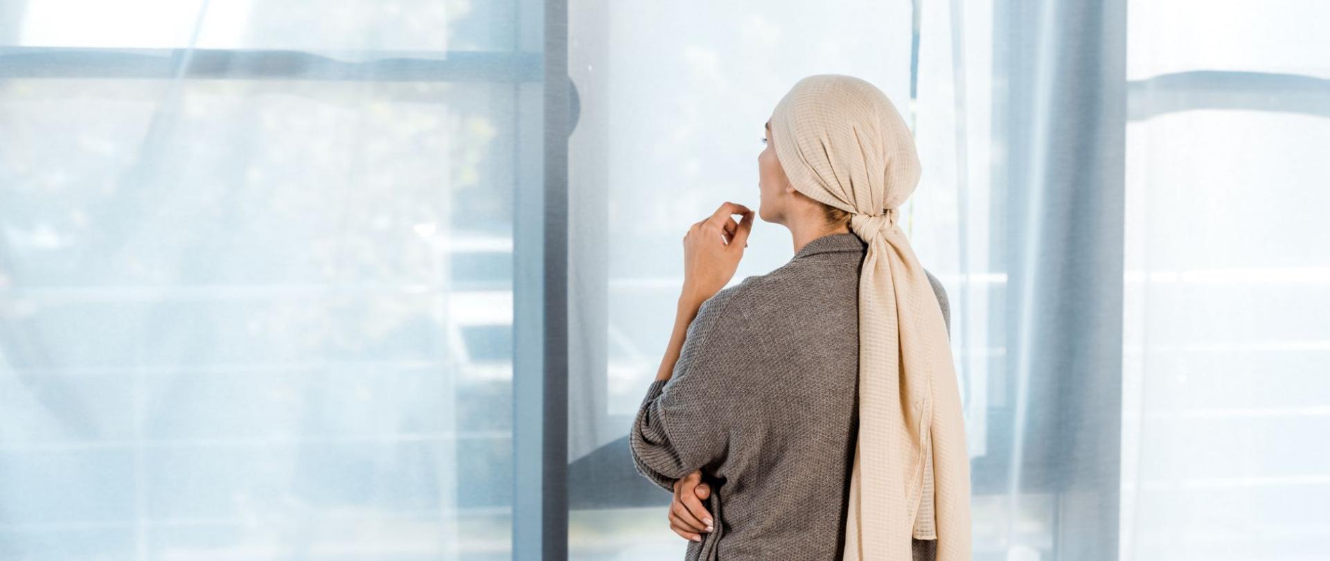 Na zdjęciu jest kobieta w chuście na głowie, wyglądająca przez okno.