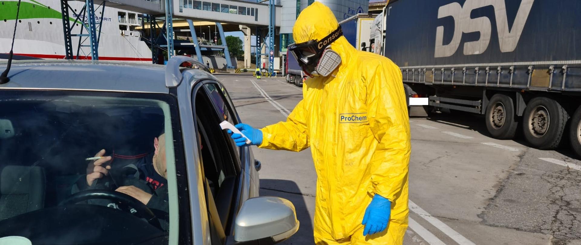 KM PSP Gdynia – podsumowanie działań z koronawirusem – na zdjęciu strażak w żółtym szczelnym kombinezonie sprawdza temperaturę kierowcy samochodu. W tle terminal portu.