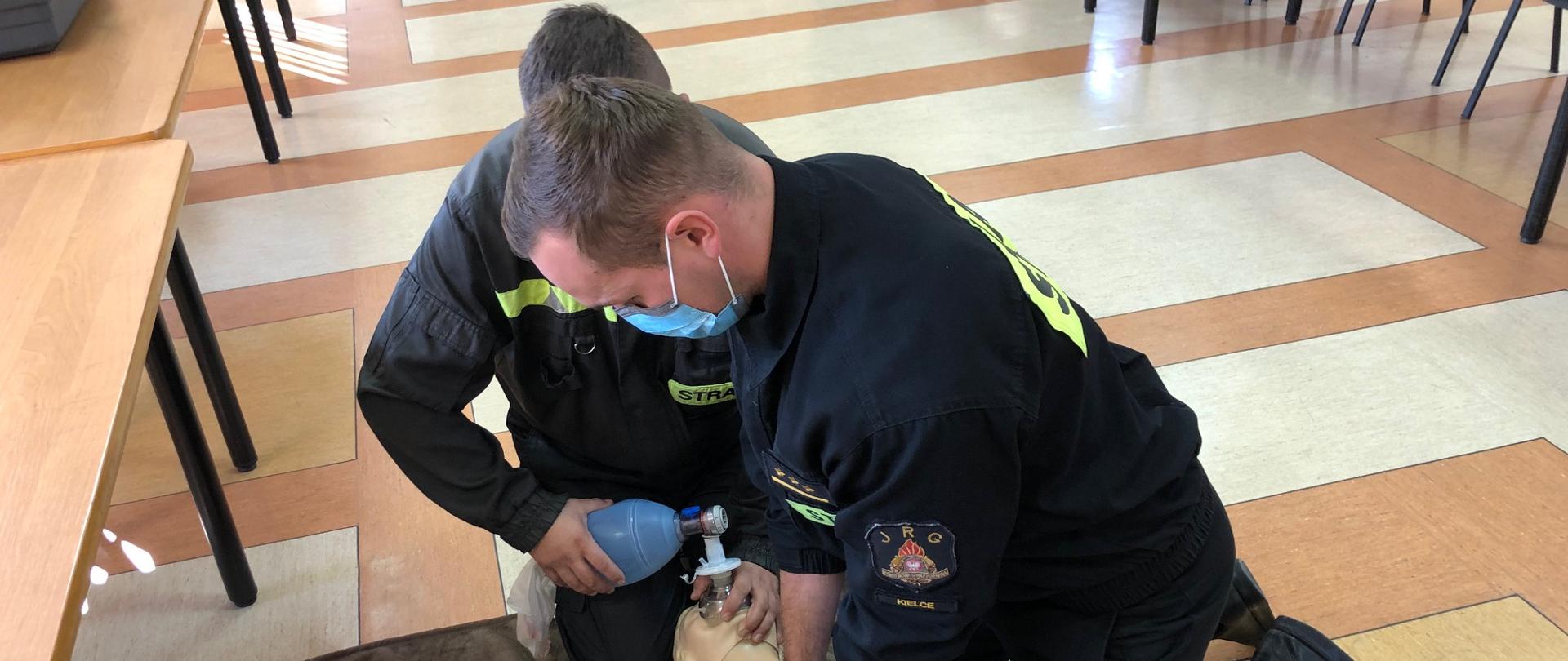 Na pierwszym zdjęciu strażacy-ratownicy wykonują symulację resuscytacji krążeniowo-oddechowej u osoby dorosłej. Jeden z ratowników wykonuje 30 uciśnięć klatki piersiowej, natomiast drugi wykonuje 2 oddechy ratownicze za pomocą worka samorozprężalnego.