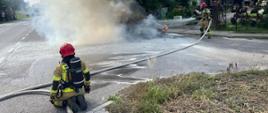 Na zdjęciu strażak gaszący palący się samochód, oraz drugi strażak klęczący przy rozdzielaczu linii gaśniczej
