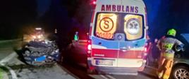 Zdjęcie przedstawia uszkodzone pojazdy biorące udział w zdarzeniu, które stoją na drodze. Na jezdni stoi również karetka pogotowia ratunkowego.