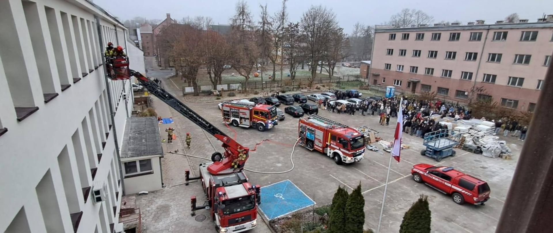 Zdjęcie z robione z góry, na szkolny plac. Na dole na placu stoją cztery samochody strażackie, samochody osobowe oraz grupa młodzieży ze szkoły. Trwają ćwiczenia strażackie, rozłożona drabina mechaniczna oraz sprzęt strażacki.