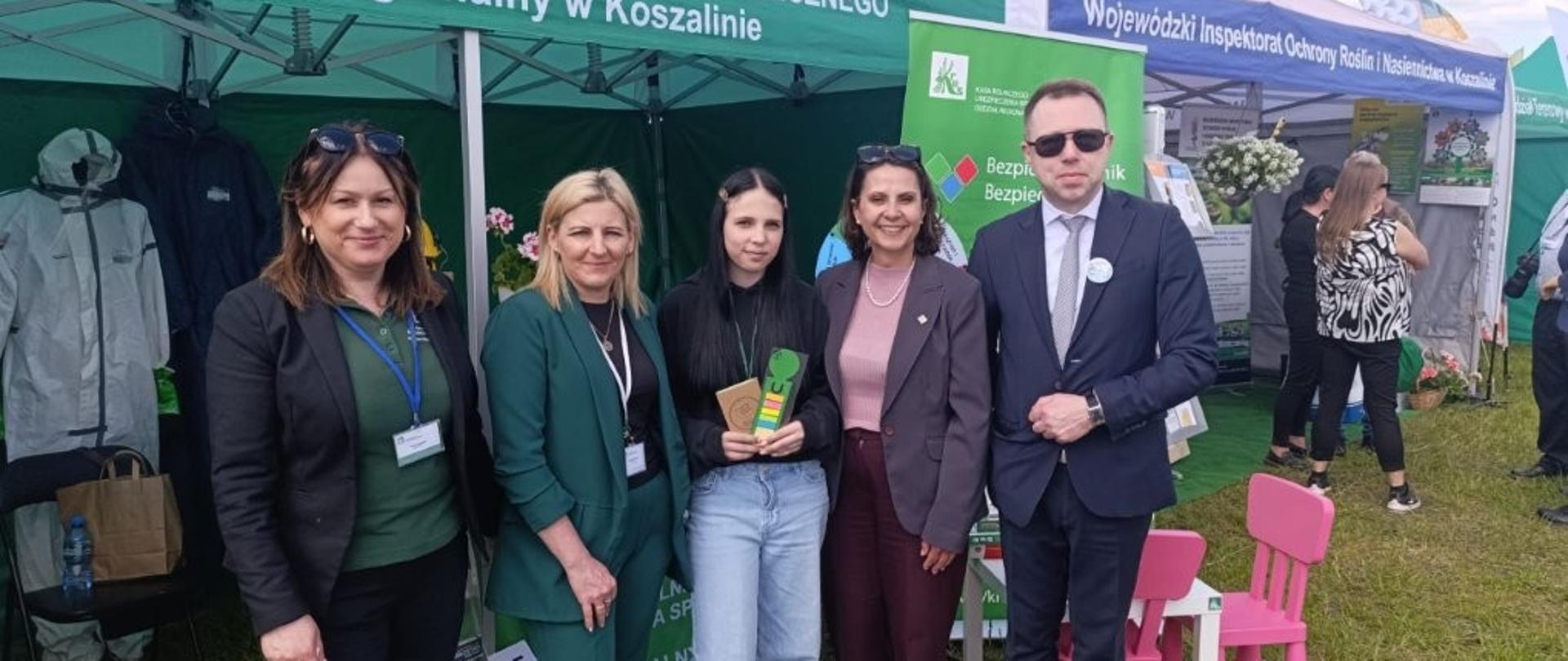 Cztery kobiety i mężczyzna pozują do zdjęcia przed zielonym namiotem KRUS. jedna z kobiet trzyma w ręce materiały promocyjne KRUS, zakładkę do książki i notatnik. Z tyłu na stoisku strój ochronny do prac rolniczych. Po prawej stoisko edukacyjne dla dzieci.