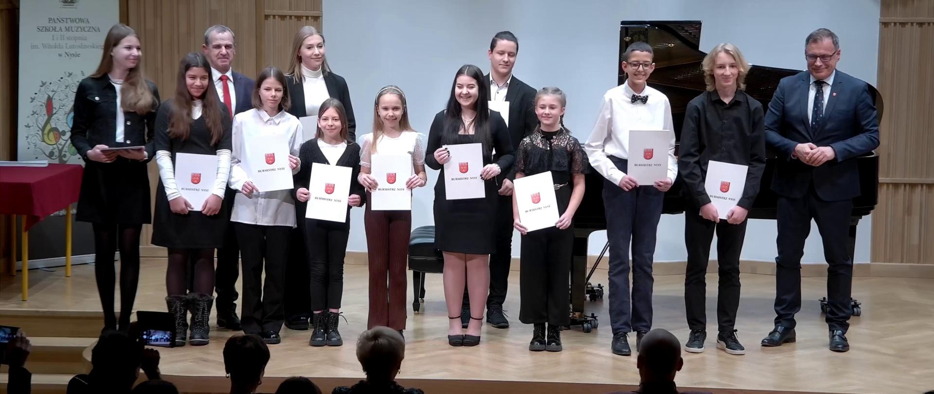 Zdjęcie. Na estradzie, przed słuchaczami, stoją laureaci stypendiów Burmistrza Nysy - uczniowie szkół w Gminie Nysa. Trzymają dyplomy. Towarzyszą im Burmistrz i Przewodniczący Rady Miejskiej.