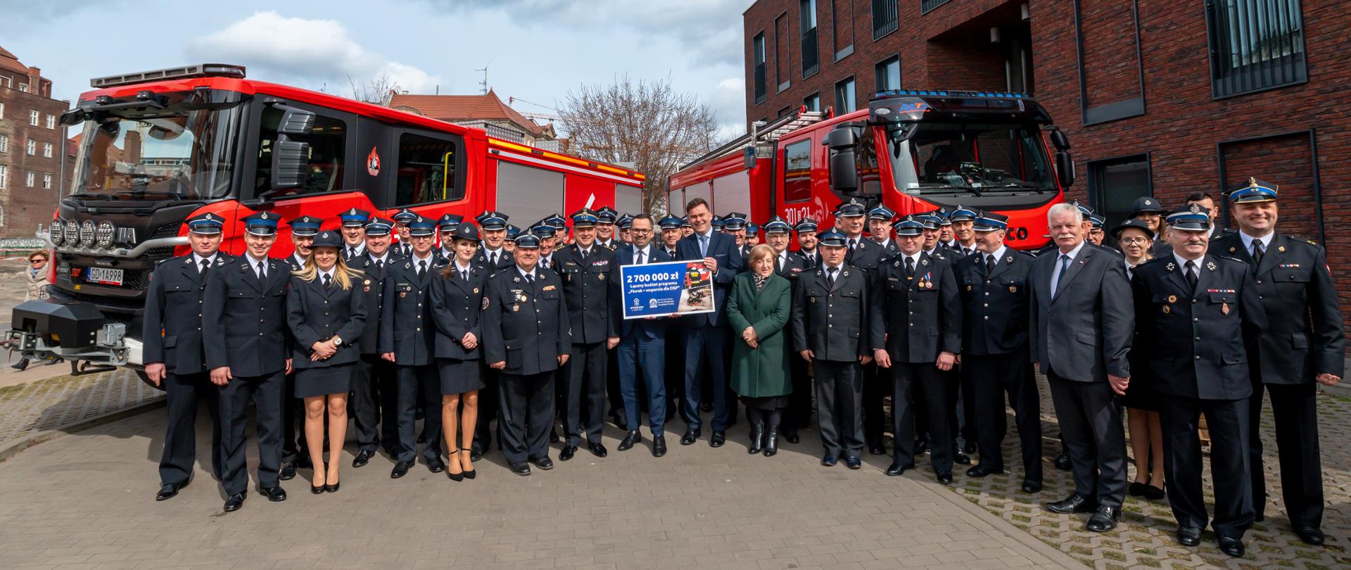 Strażacy ochotniczych straży pożarnych, zaproszeni goście stoją na tle samochodów gaśniczych.