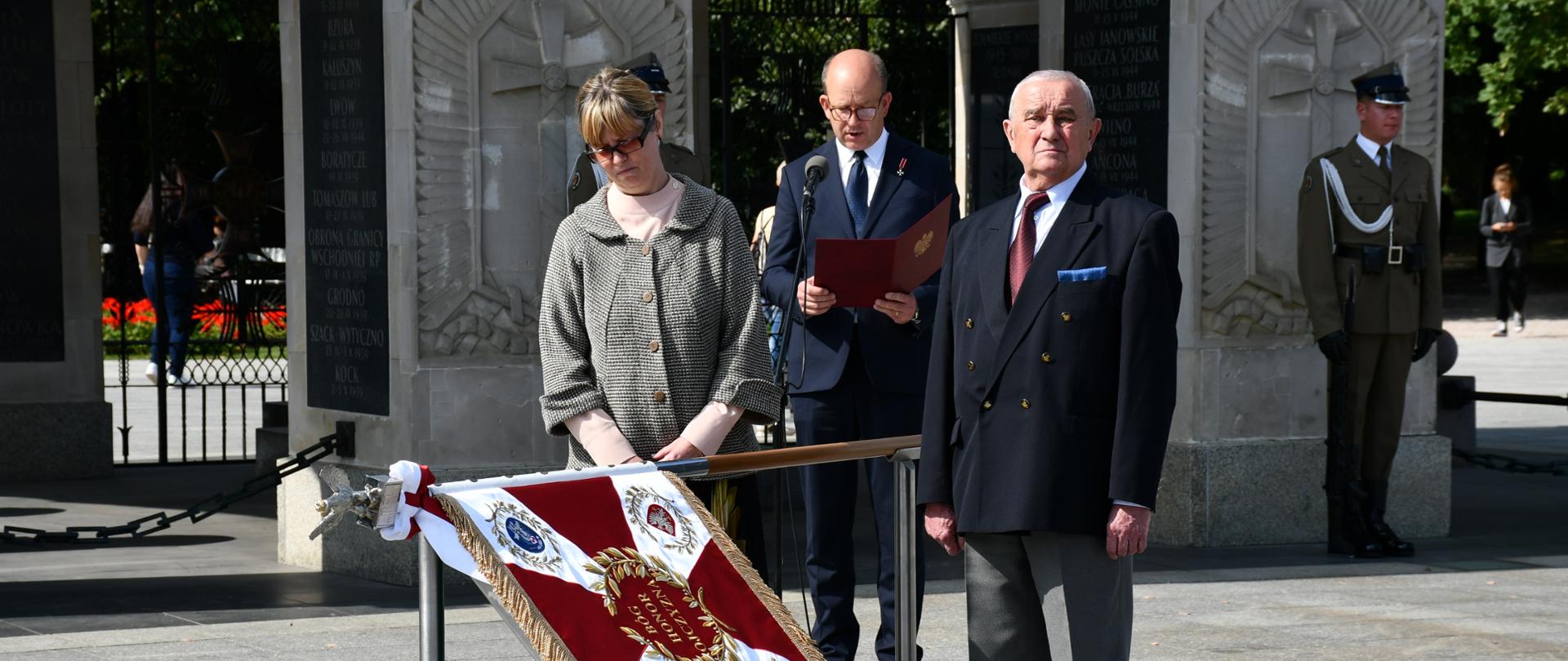 Na zdjęciu znajduje się sztandar, za nimi trzy osoby