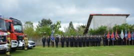 Pododdziały w szeregu w trakcie uroczystości. Sztandary jednostek OSP oraz samochody pożarnicze po lewej stronie zdjęcia.