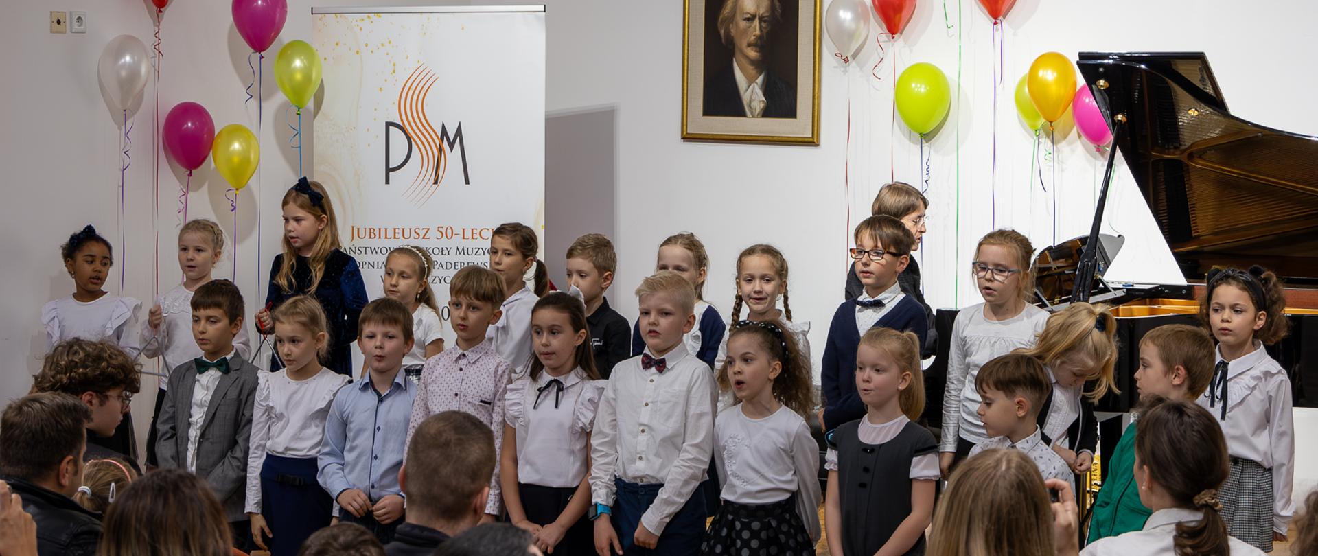 Pierwszoklasiści śpiewają wspólnie na scenie PSM w Głubczycach podczas uroczystości pasowania na ucznia. Przy fortepianie pani Małgorzata Meyse. W dolnej części zdjęcia widoczna publiczność. Scena na uroczystość przyozdobiona została kolorowymi balonikami.