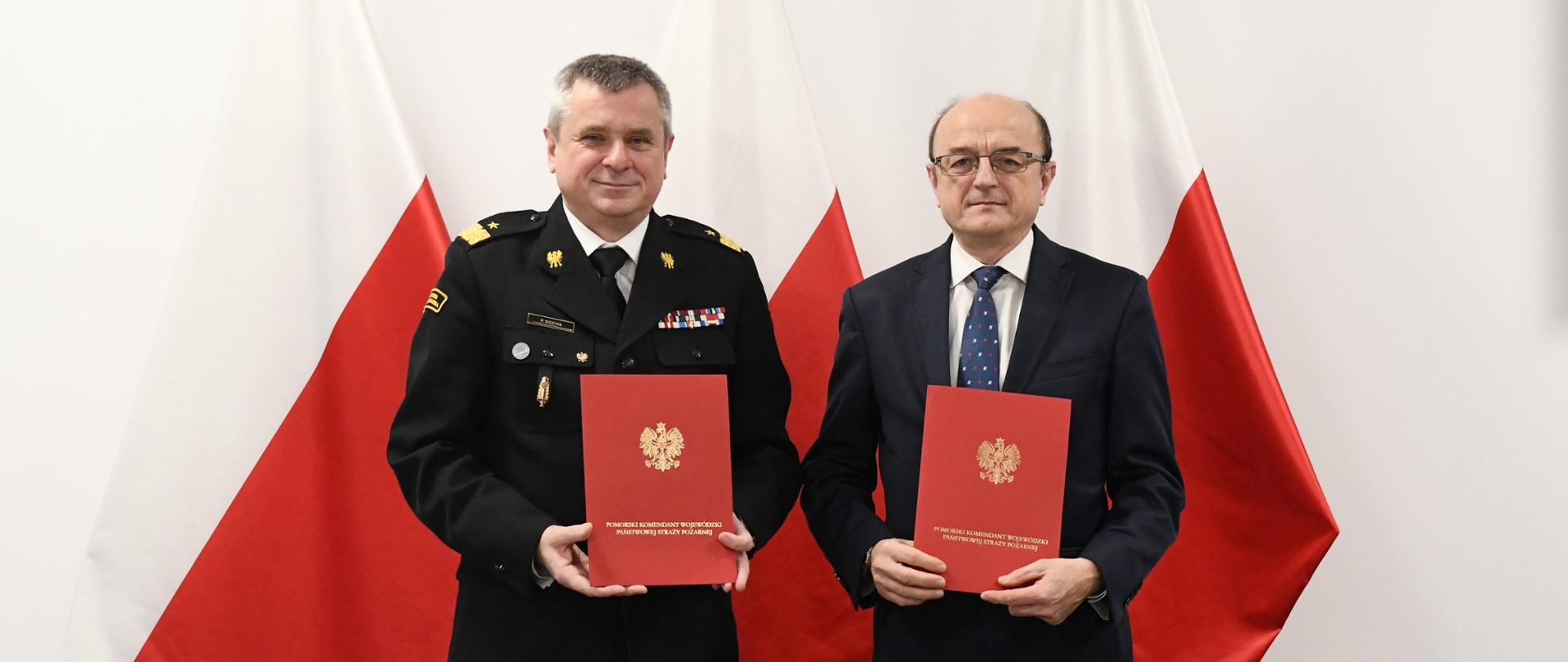 Strażak oraz mężczyzna trzymają czerwone teczki za nimi są flagi Polski.