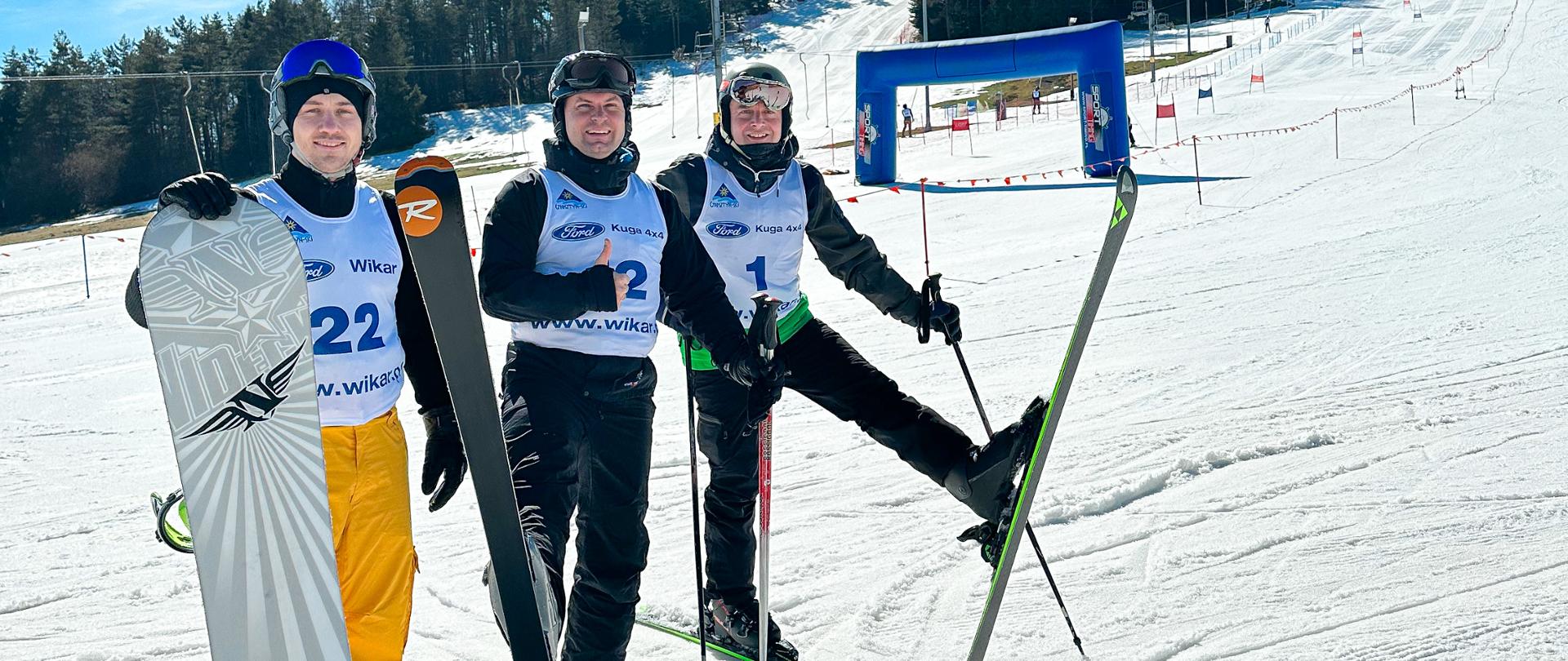 Zdjęcie przedstawia trzech zawodników ubranych w koszulki z numerami startowymi. Dwóch z założonymi nartami oraz trzeci trzymający deskę snowboardową. Zawodnicy pozują na śniegu, w tle widać tor slalomu narciarskiego/snowboardowego oraz metę.