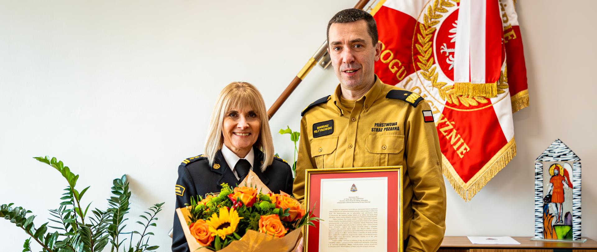 Na zdjęciu widać strażaka w umundurowaniu wyjściowym. W tle sztandar strażay pożarnej. Strażak trzyma dyplom z gratulacjami za wieloletnią służbę.