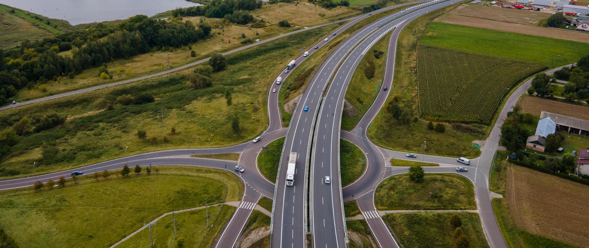 Widok z lotu ptaka na dwujezdniową drogę ekspresową z węzłem drogowym. Jezdnie rozdzielone pasem zieleni, po bokach drogi serwisowe i pola uprawne. W tle widoczne zbiorniki wodne, zabudowa miejscowości oraz rozległy krajobraz rolniczy pod pochmurnym niebem.