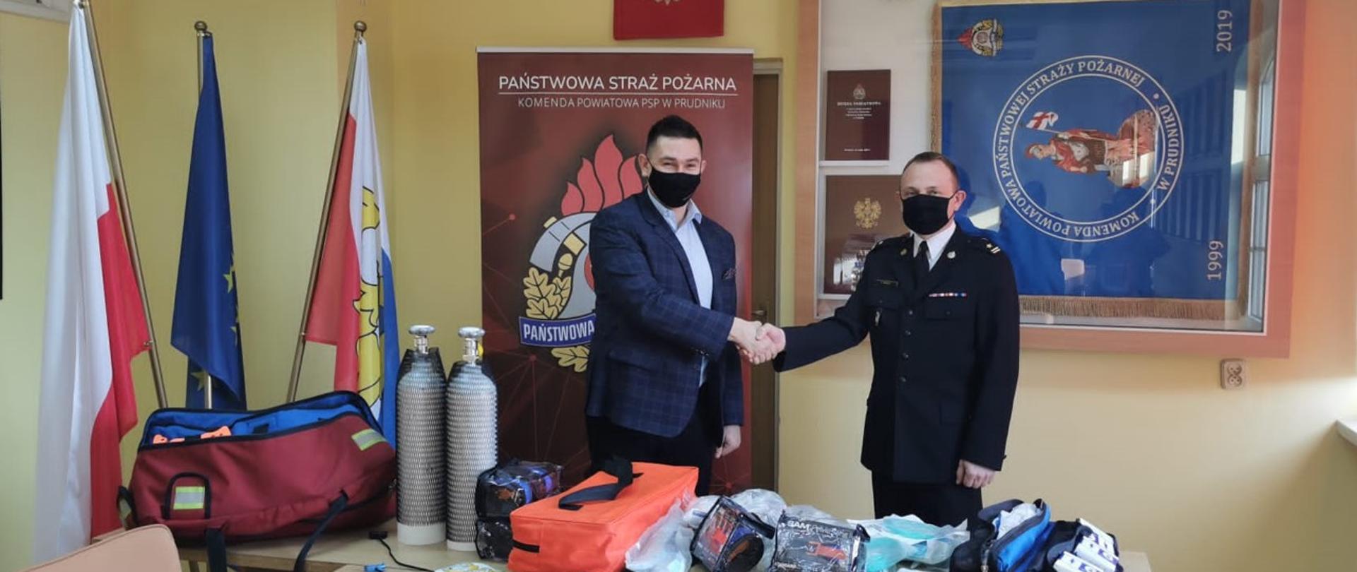 Zdjęcie przedstawia przekazanie sprzętu do ratownictwa medycznego dla KP PSP Prudnik. Na zdjęciu stoją Burmistrz Głogówka oraz Komendant Powiatowy PSP w Prudnik. W tle po lewej stronie zdjęcia flagi, na środku Godło Rzeczpospolitej Polskiej i logo PSP, po prawej stronie gablota ze sztandarem KP PSP Prudnik
