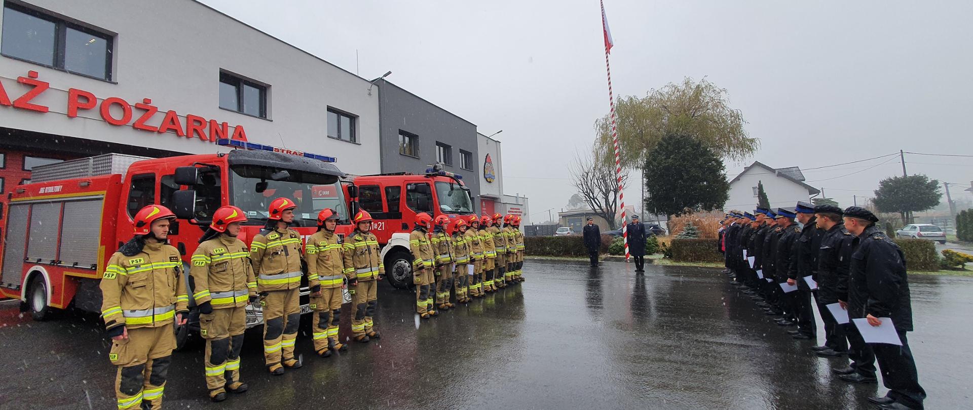 Na zdjęciu funkcjonariusze podczas uroczystej zbiórki. W tle samochody pożarnicze oraz budynek Komendy Powiatowej PSP. Pada śnieg.