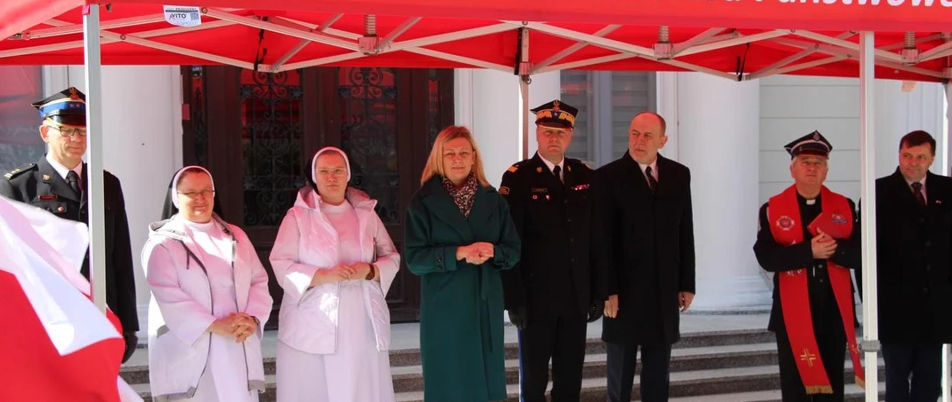 grupa ludzi w tym strażacy, ksiadz i siostry zakonne stoją pod namiotem