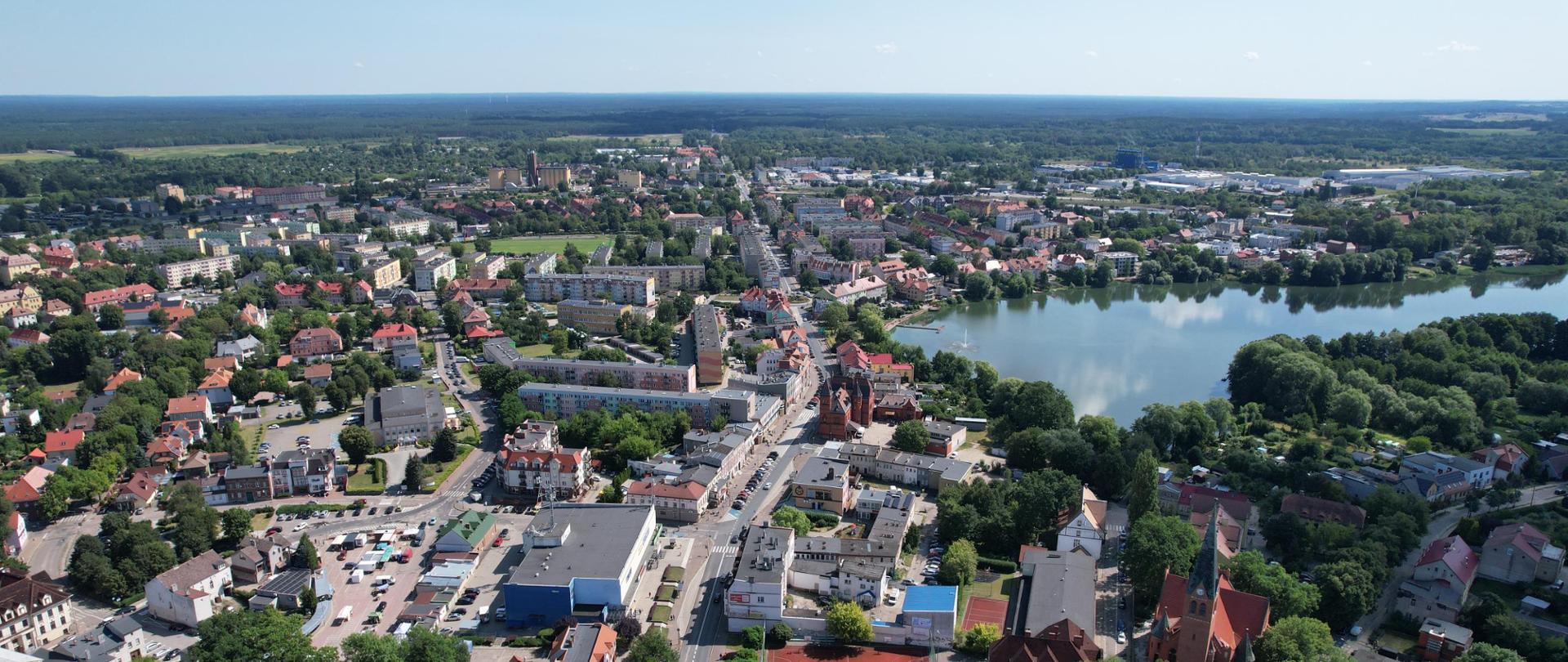 Widok z lotu ptaka na miasto. Widać różnorodne budynki, ulice oraz obszary zielone. Po prawej stronie znajduje się duże akwen wodny. Architektura obejmuje domy mieszkalne z czerwonymi dachami, większe budynki wielopiętrowe oraz ulice obsadzone drzewami. Okolica jest otoczona bujną zielenią i przestrzeniami otwartymi.