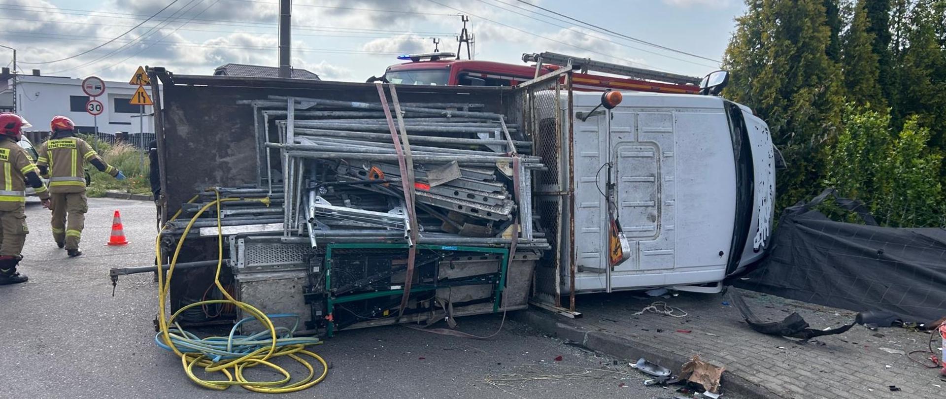 Zdjęcie przedstawia miejsce zdarzenia drogowego z udziałem samochodu dostawczego, który przewrócił się na bok. Pojazd leży na prawym boku na poboczu drogi, a jego ładunek – konstrukcje stalowe lub aluminiowe, prawdopodobnie rusztowania – częściowo się wysypał. Na miejscu obecne są jednostki straży pożarnej – widocznych dwóch strażaków w pełnym umundurowaniu ochronnym i hełmach. W tle znajduje się wóz strażacki z włączonymi światłami ostrzegawczymi. Droga została częściowo zabezpieczona pachołkami. Warunki pogodowe są dobre, a droga wydaje się być lokalna, biegnąca przez teren zabudowany. Na miejscu widoczne są również fragmenty rozbitego pojazdu oraz narzędzia i przewody.