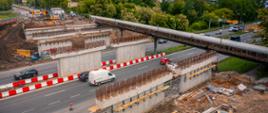 Zdjęcie przedstawia plac budowy mostu lub wiaduktu. Widać kilka betonowych konstrukcji z wystającym zbrojeniem, co świadczy o trwających pracach. Przez środek kadru przebiega droga, po której poruszają się samochody i biały van. Nad drogą znajduje się kładka dla pieszych. Po prawej stronie widoczna jest żółta koparka, a po lewej – pracownicy w odzieży ostrzegawczej w pobliżu maszyn. W tle znajdują się drzewa i tereny zielone.