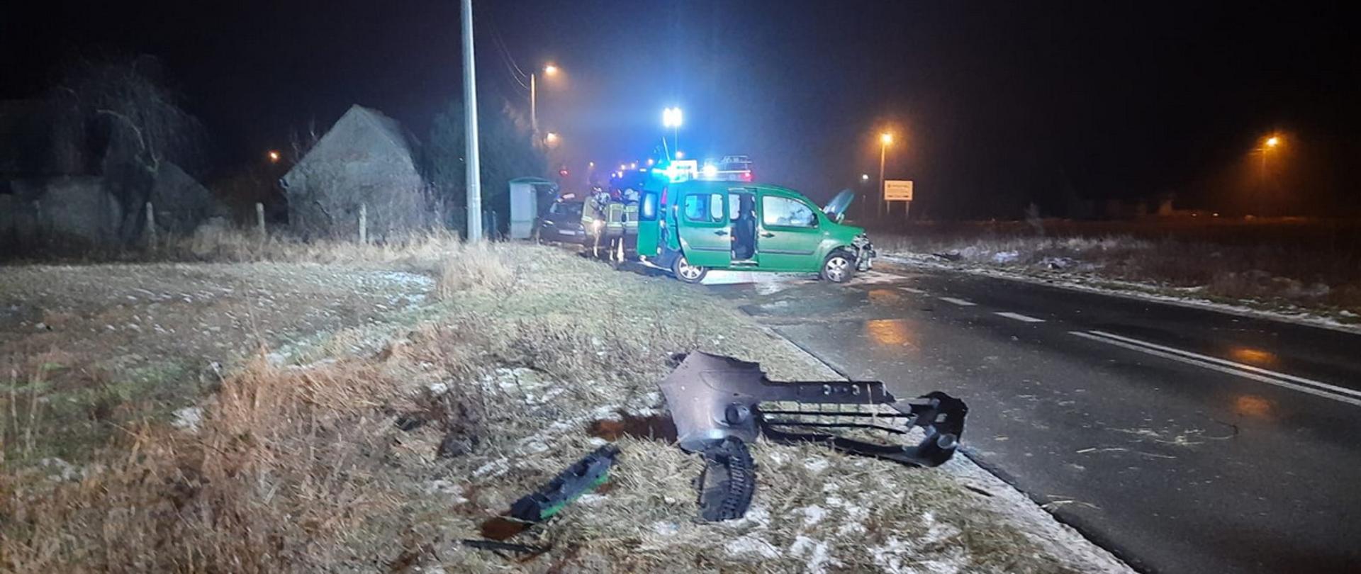 Samochód osobowy z uszkodzonym przodem stoi na środku drogi. Na pierwszym planie zniszczone elementy karoserii pojazdy, w oddali widoczne zastępy straży pożarnej.