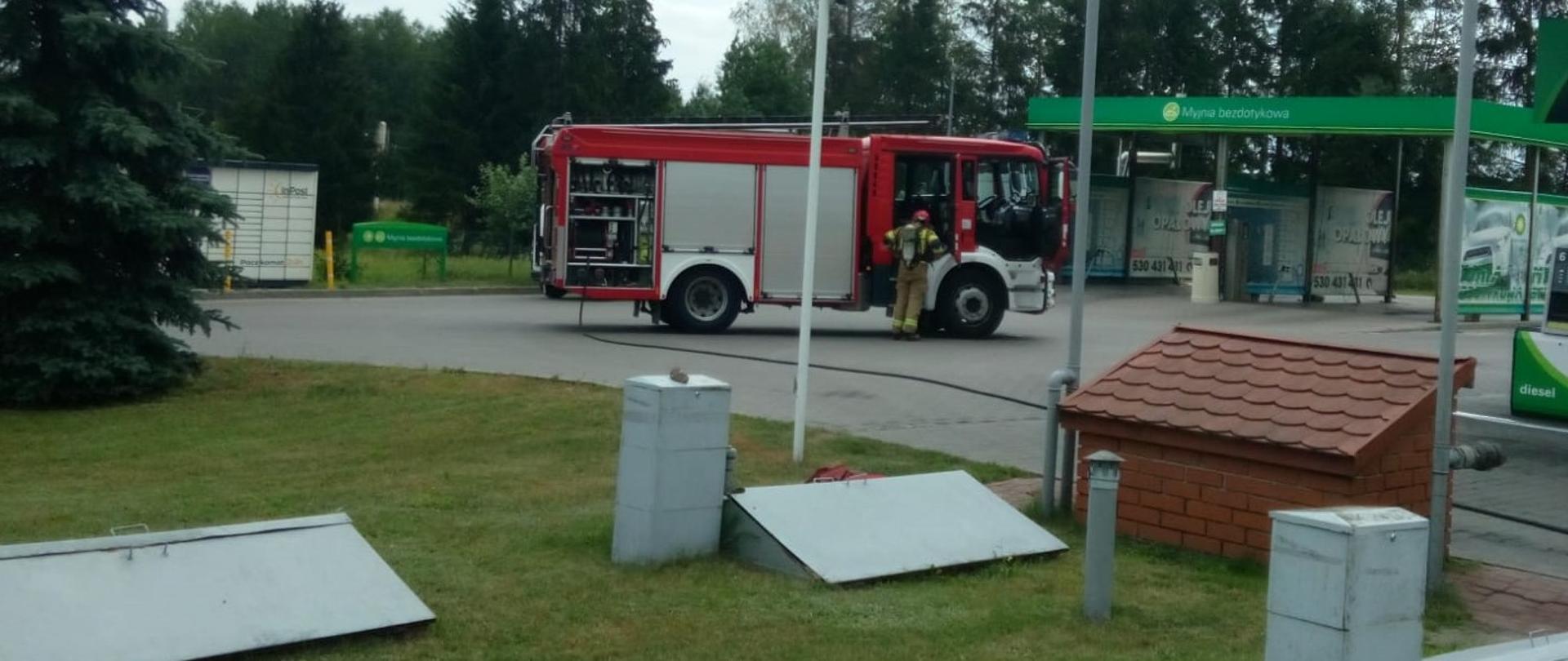 Zdjęcie przedstawia stację benzynową gdzie doszło do zadymienia. Widać zaparkowany samochód pożarniczy do którego podchodzi zabezpieczony w aparat dróg oddechowych strażak. Od samochodu rozłożona jest linia gaśnicza, a na pierwszym planie metalowe pokrywy komór dystrybutorów.
