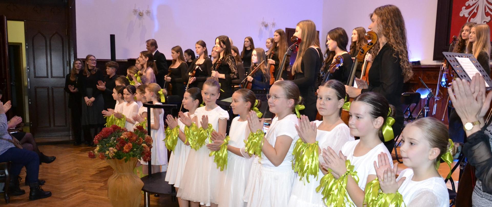 Uczniowie i nauczyciele na scenie podczas koncertu Vivaldiego: na pierwszym planie dzieci w białych strojach wykonują układ taneczny, w tle orkiestra kameralna z wiolinistkami i wiolonczelami oraz solistki i soliści prezentujący wybrane części z Czterech pór roku