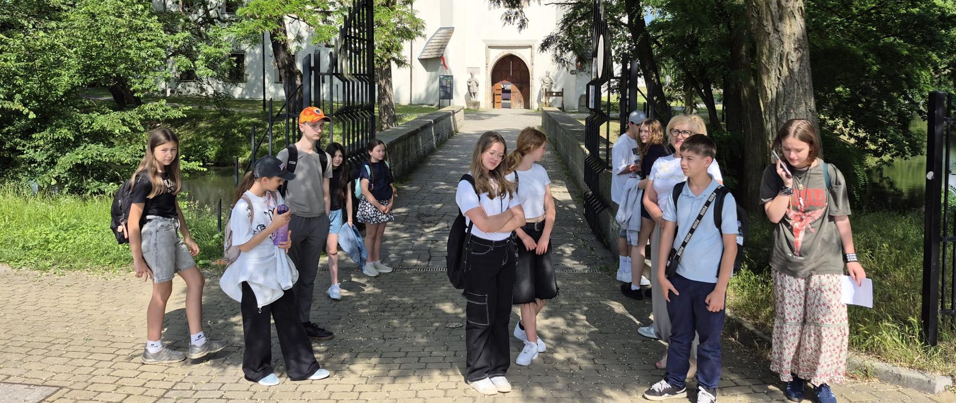 Zdjęcie w pionie wykonane w słoneczny dzień. Na pierwszym planie grupa ok. 12 młodych osób i jedna dorosła kobieta stoi na brukowanej ścieżce. Są ubrani letnio, większość ma plecaki. Dwie dziewczynki na środku patrzą w obiektyw, jedna ma założone ręce. Chłopak po lewej ma pomarańczową czapkę, dziewczyna po prawej rozmawia przez telefon.
Za nimi wąski kamienny most z czarnymi, ozdobnymi barierkami prowadzi nad wodą. Na końcu mostu stoi biały, zabytkowy budynek z czerwonym dachem i dużą, łukową, drewnianą bramą. Po obu stronach wejścia stoją kamienne figury. Całość otaczają gęste, zielone drzewa rzucające cień na ścieżkę.