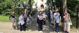 Zdjęcie w pionie wykonane w słoneczny dzień. Na pierwszym planie grupa ok. 12 młodych osób i jedna dorosła kobieta stoi na brukowanej ścieżce. Są ubrani letnio, większość ma plecaki. Dwie dziewczynki na środku patrzą w obiektyw, jedna ma założone ręce. Chłopak po lewej ma pomarańczową czapkę, dziewczyna po prawej rozmawia przez telefon.
Za nimi wąski kamienny most z czarnymi, ozdobnymi barierkami prowadzi nad wodą. Na końcu mostu stoi biały, zabytkowy budynek z czerwonym dachem i dużą, łukową, drewnianą bramą. Po obu stronach wejścia stoją kamienne figury. Całość otaczają gęste, zielone drzewa rzucające cień na ścieżkę.